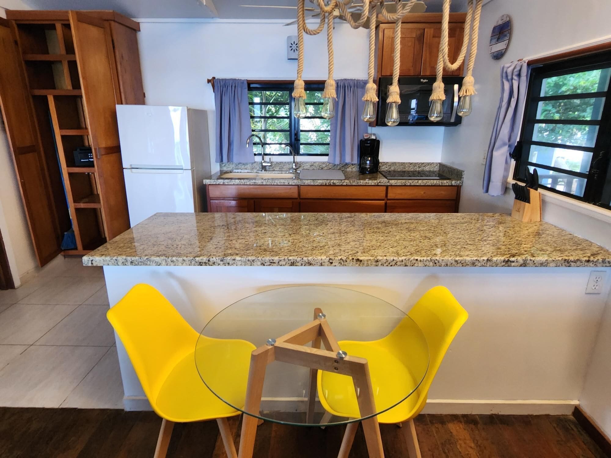 Uma cozinha com bancadas de granito, geladeira branca, armários de madeira, janela com cortinas e uma cadeira amarela em uma mesa de vidro.