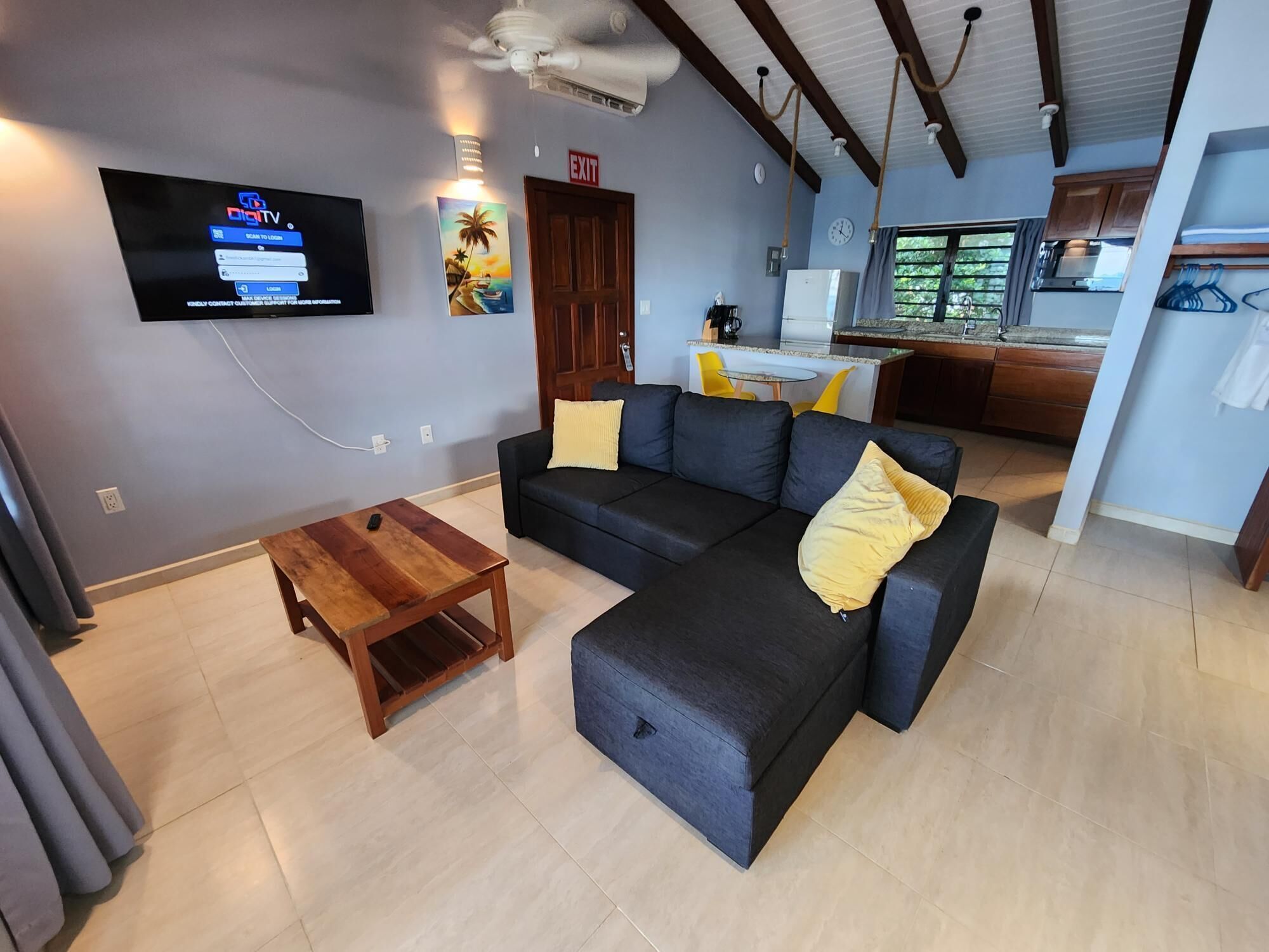 A living room with a dark sofa, a wooden coffee table, a flat-screen TV mounted on the wall, and a kitchen area visible through an open door.