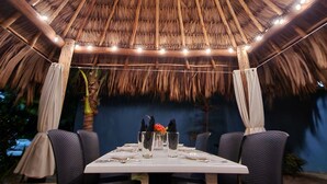 An outdoor dining area with a thatched roof, a table set for two, and black chairs.