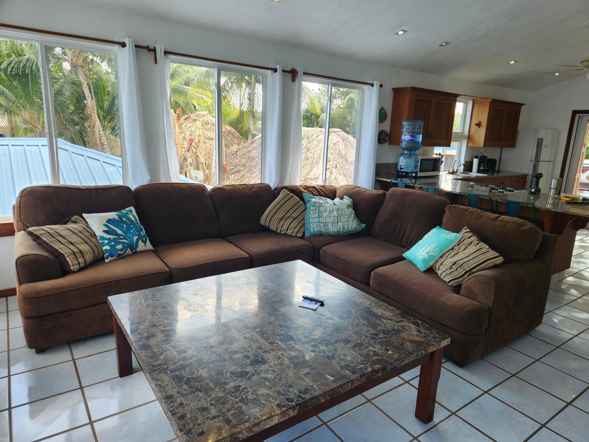 Una sala de estar con un sofá seccional marrón, una mesa de centro de mármol y ventanas grandes que dan a una vista de palmeras y una estructura de techo de paja.