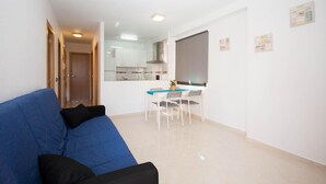 Una sala de estar moderna con un sofá azul, un comedor con una mesa y sillas de vidrio, y una cocina al fondo.