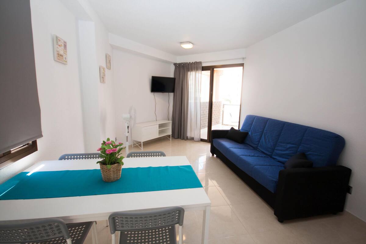 A living room with a white table, blue sofa, and a TV mounted on the wall.