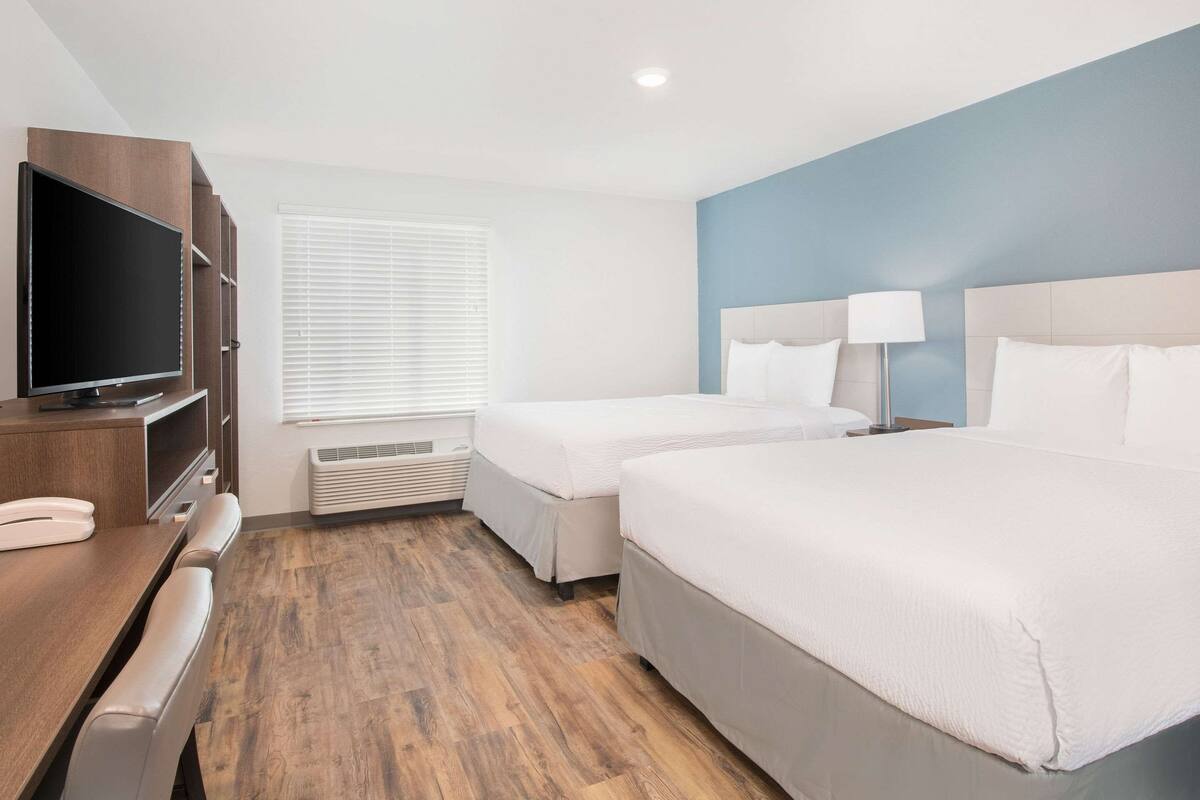 A hotel room with two beds, a TV, a desk, and a window with blinds.