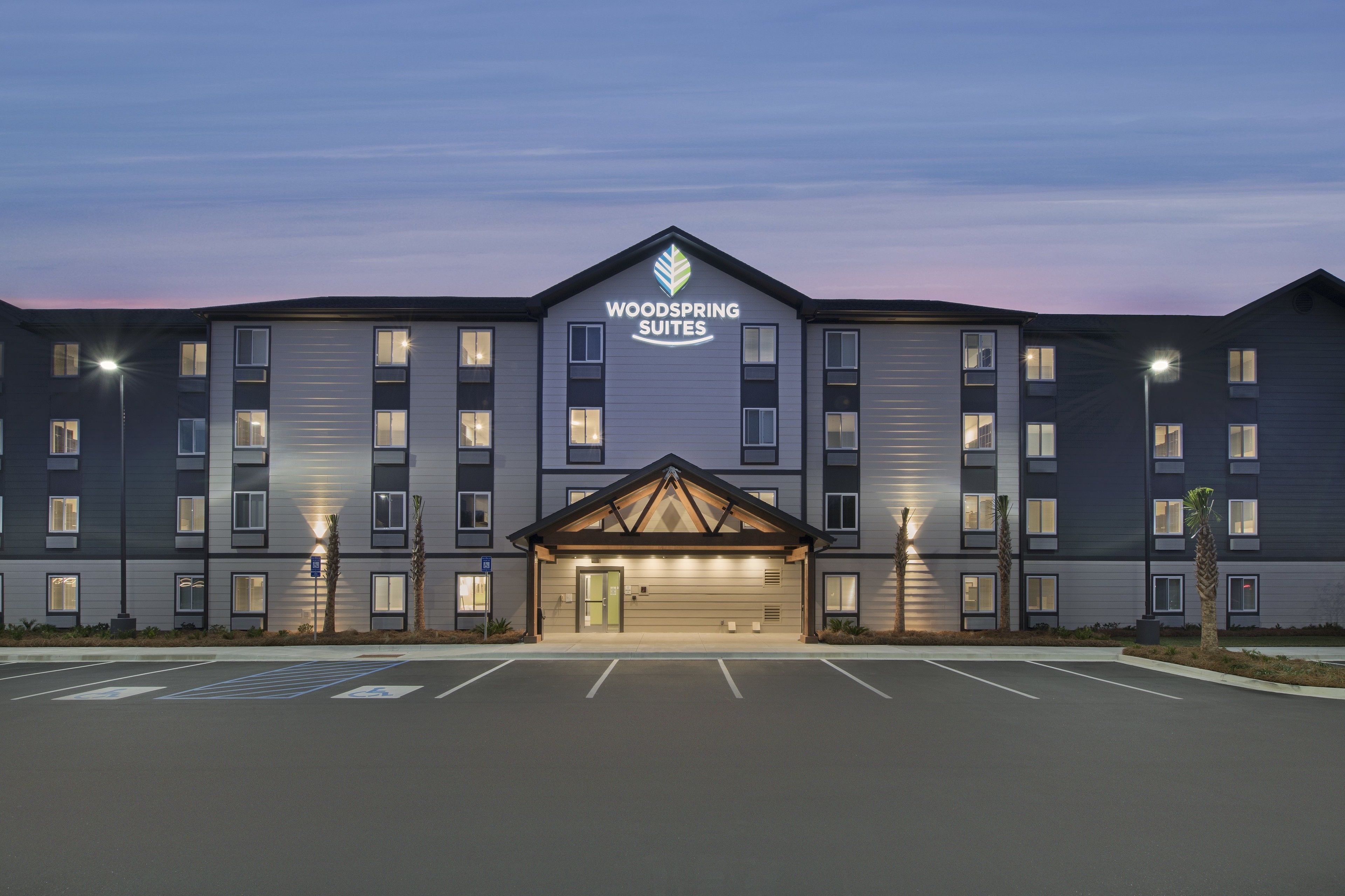 A multi-story hotel with a prominent "Woodspring Suites" sign, a central entrance with a ramp, and a parking lot with marked handicapped spaces.