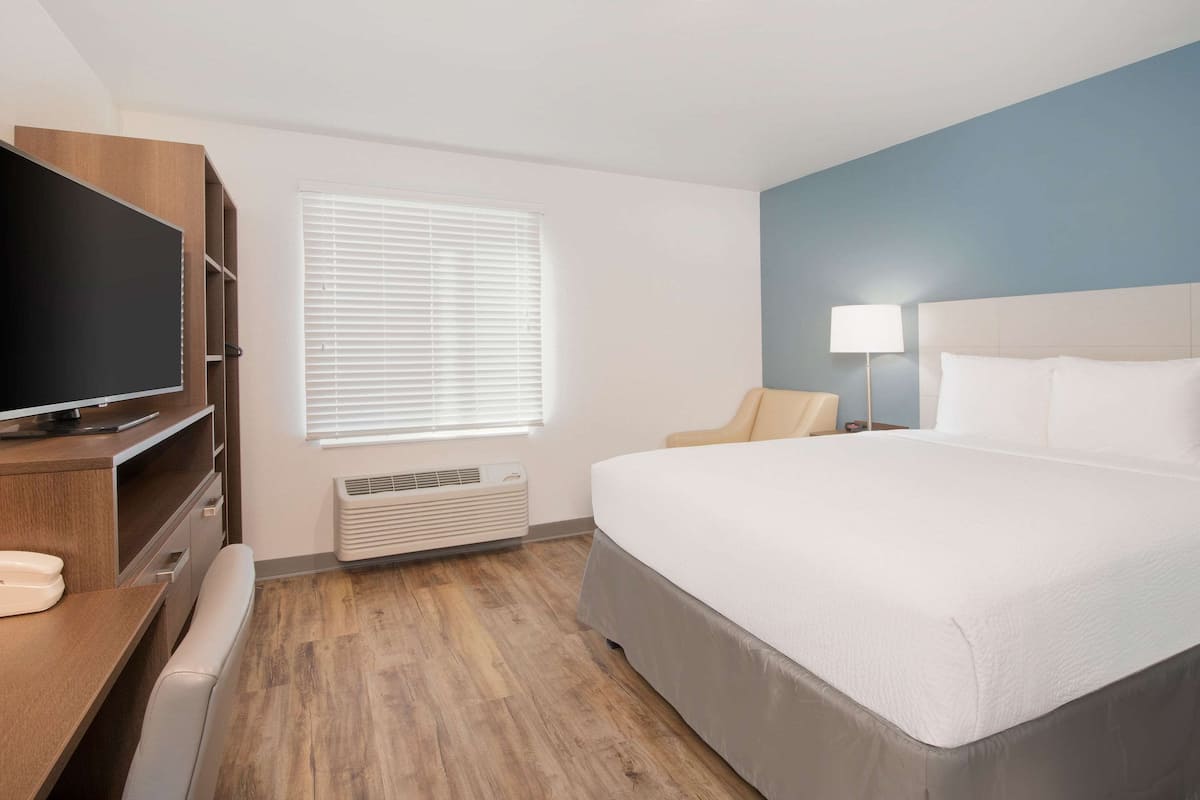 A hotel room with a large bed, a desk with a flat-screen TV, a chair, a bedside lamp, and a window with blinds.