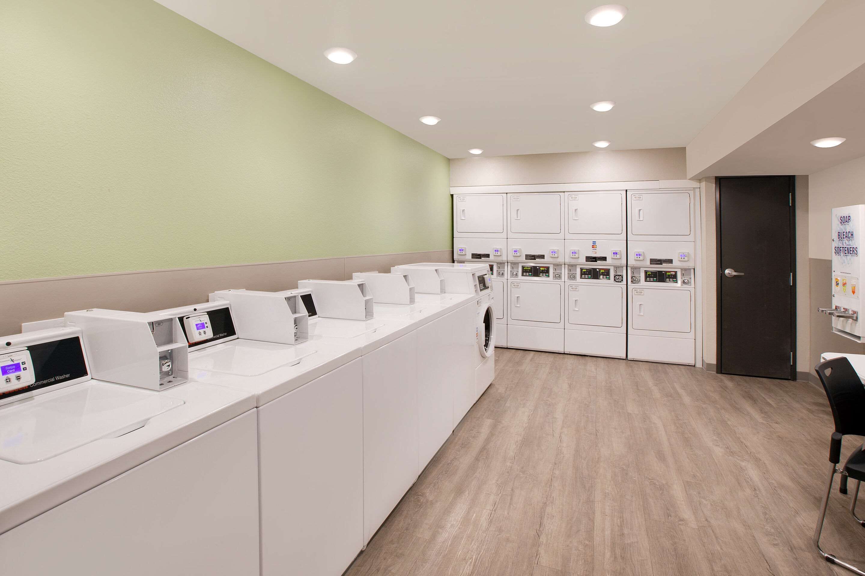 A modern laundry facility with multiple washing machines and dryers, a wooden floor, and a green accent wall.