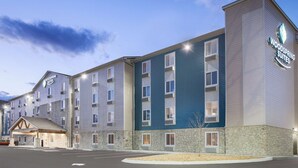 Een meerlaags hotel met een bord waarop 'Woodspring Suites' staat. Het heeft een gevel van natuursteen, meerdere balkons en een parkeerplaats ervoor.
