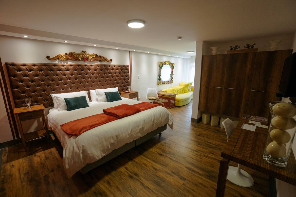 A hotel room with a large bed, wooden flooring, a wooden headboard, a mirror, a wardrobe, and a desk with a chair.