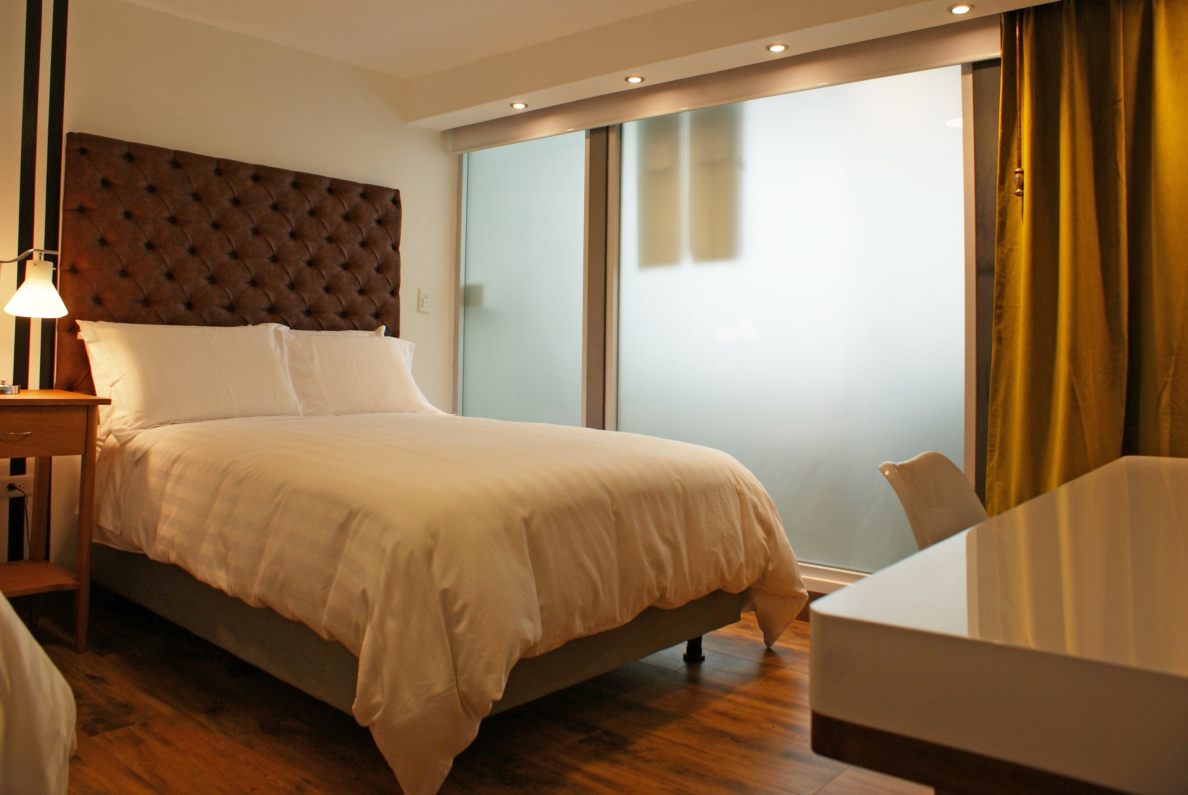 Habitación de hotel con una cama grande, una mesita de noche con lámpara, una puerta de cristal que da a un balcón y vistas al exterior.
