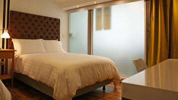 Habitación de hotel con una cama grande, una mesita de noche con lámpara, una puerta de cristal que da a un balcón y vistas al exterior.