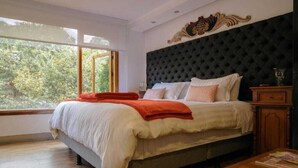 Een slaapkamer met een groot bed, een donker gewatteerd hoofdbord en uitzicht op bomen door een groot raam.