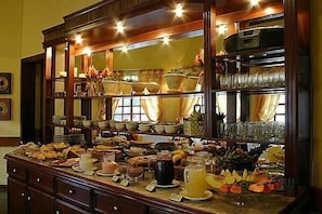 Buffet com diversos alimentos e bebidas, disposto num balcão com um armário de madeira ao fundo.