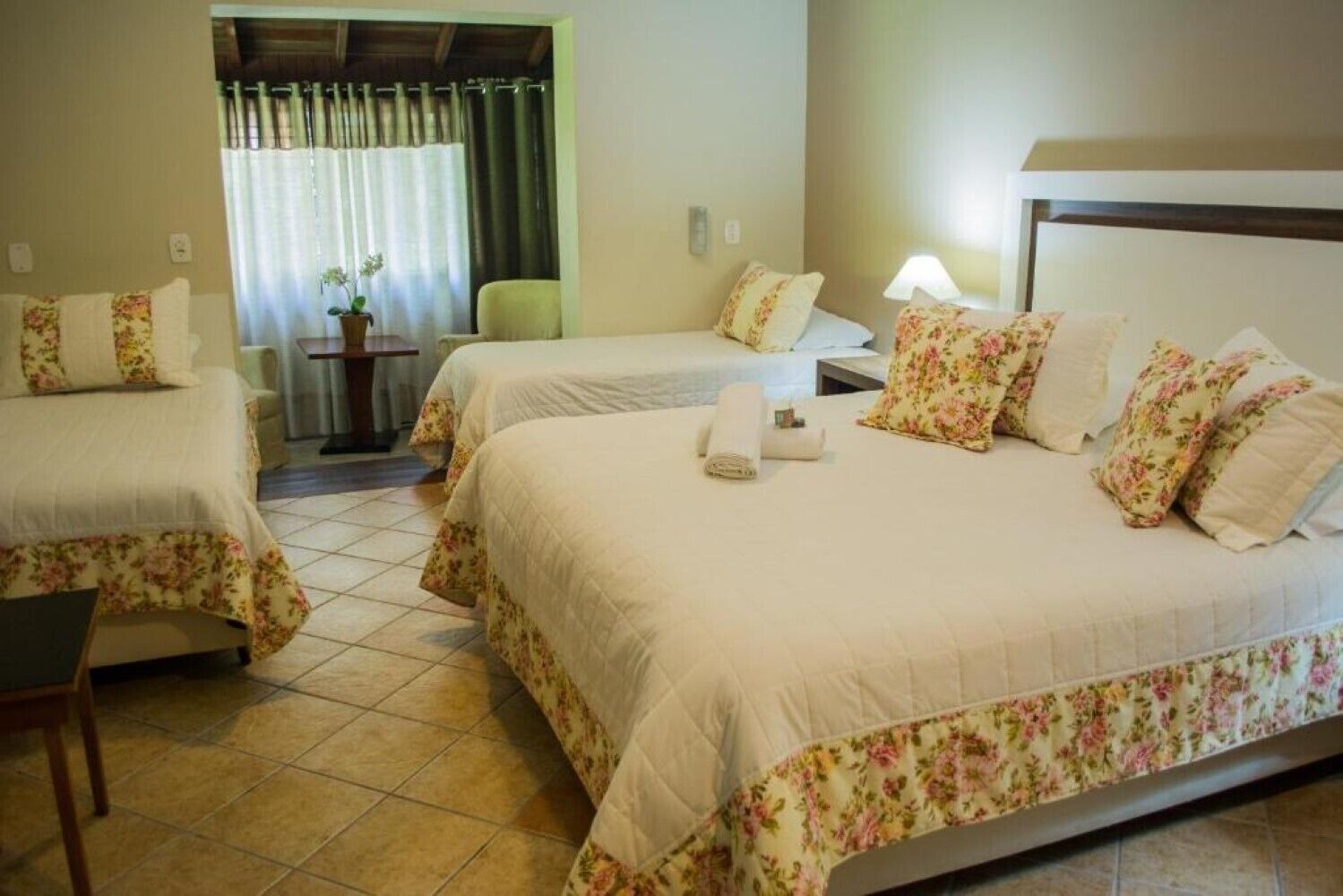 A hotel room with two beds, each with floral bedding and pillows, a small table with a lamp, and a view of a garden through a window with curtains.