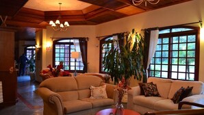 A cozy living room with two sofas, a round coffee table, and a vase with flowers. There are large windows with curtains, a chandelier, and a person in the background. - Pousada do Bosque (Canela)