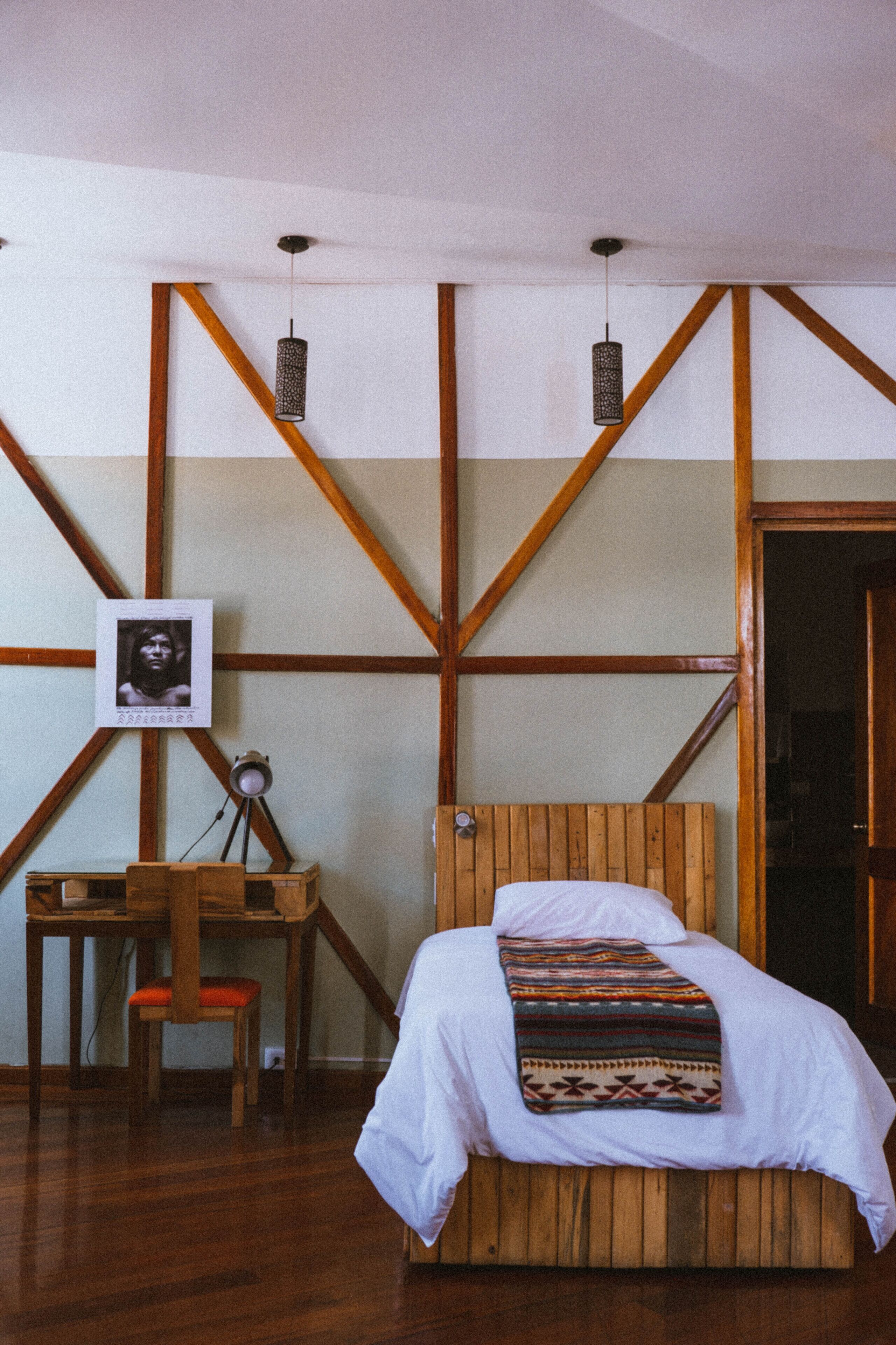 Una habitación con una cama de madera, un escritorio y una silla. Hay un cuadro en la pared y una puerta a la derecha.