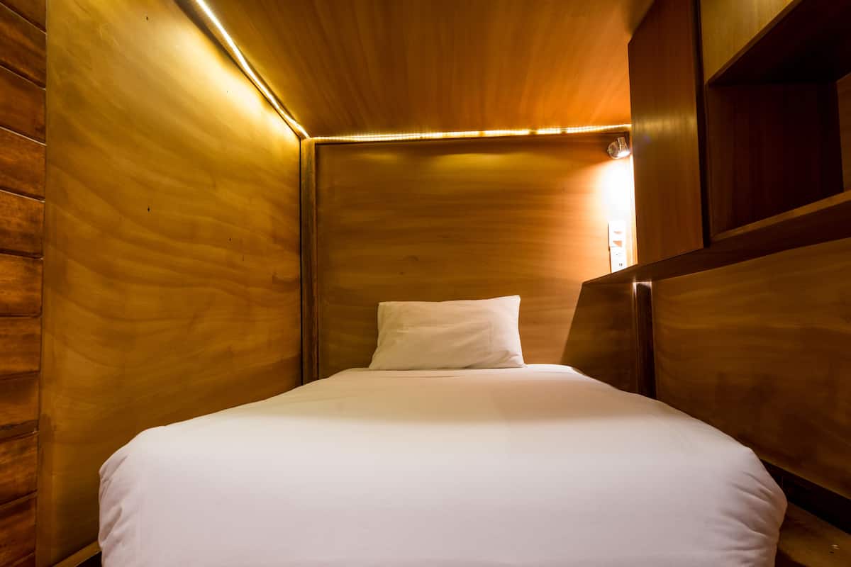 A single bed with a white mattress and pillow in a wooden room with a sloped ceiling and built-in shelving.