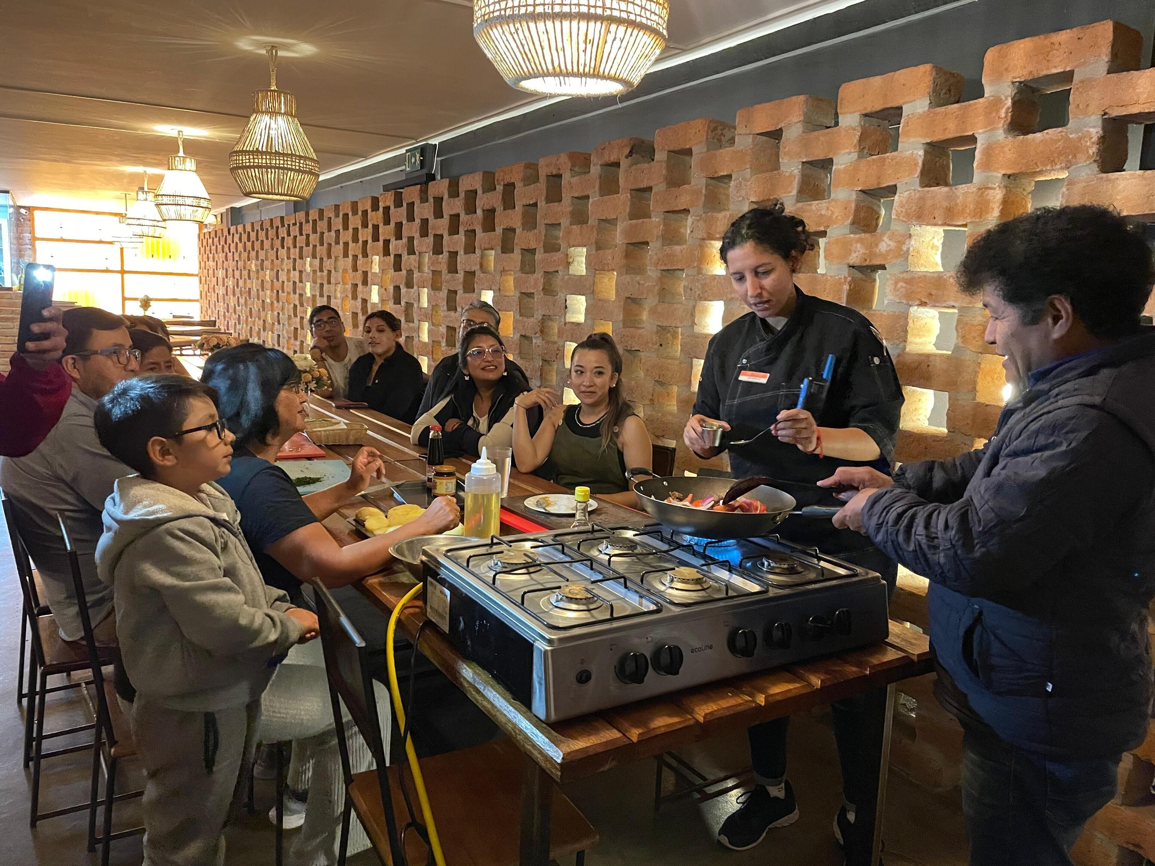 A group of people gathered around a cooking station with a gas stove, in a room with brick walls and hanging lights.