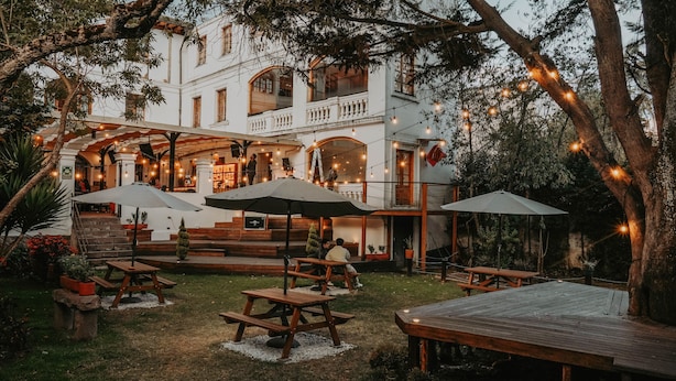 An outdoor patio with wooden tables and umbrellas, surrounded by trees and a white building with balconies. - Viajero Quito Hostel (Quito)
