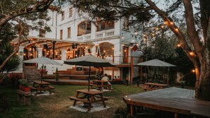 An outdoor patio with wooden tables and umbrellas, surrounded by trees and a white building with balconies. - Viajero Quito Hostel (Quito)