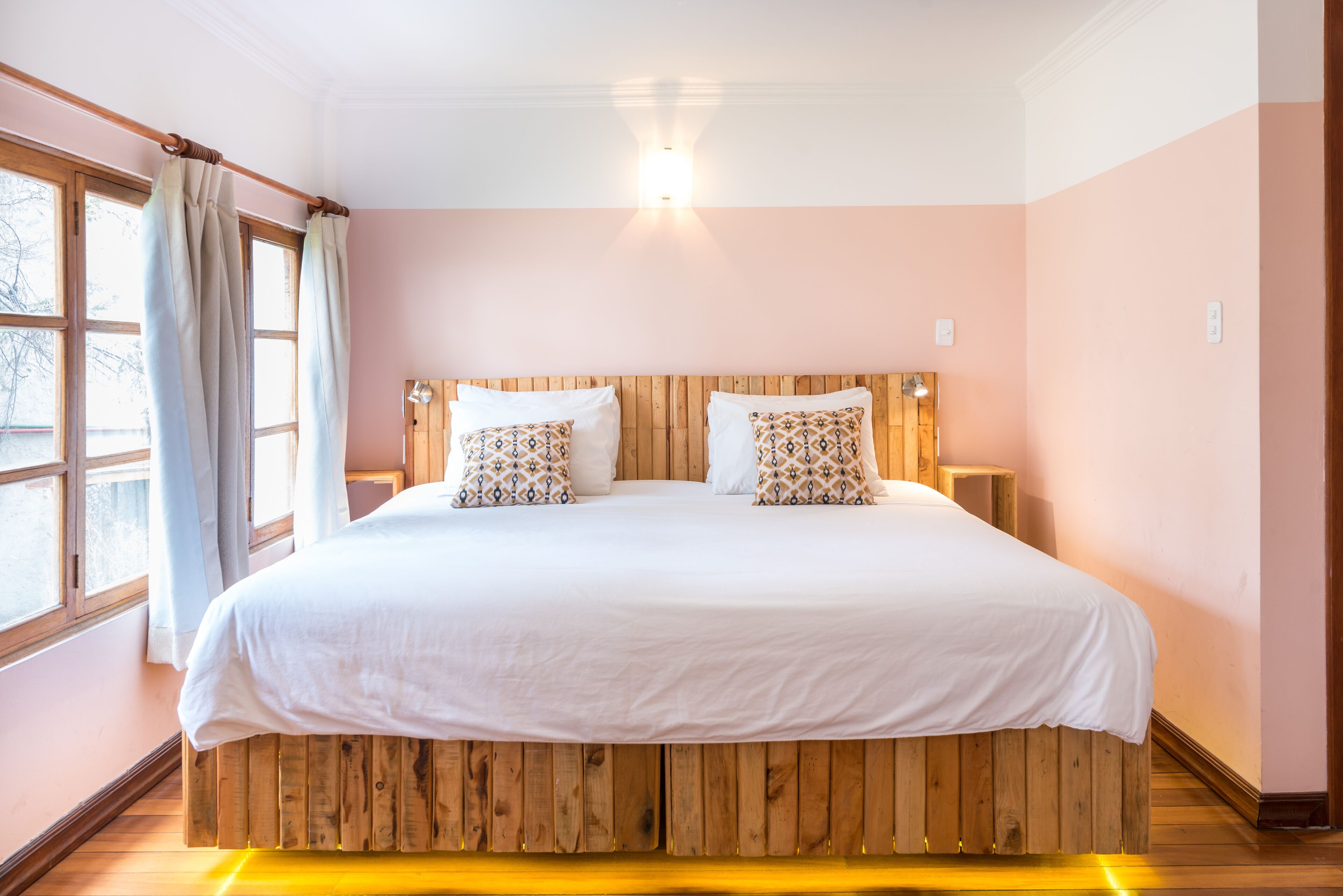 A bedroom with a wooden headboard, two white bedsheets, patterned pillows, a bedside table with a lamp, and a window with curtains.