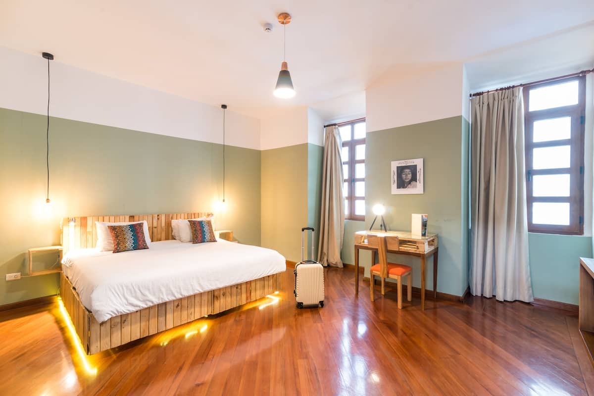 A hotel room with a wooden bed, a desk, a chair, a suitcase, and a window with curtains.