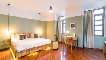 Habitación de hotel con cama de madera, escritorio, silla, maleta y ventana con cortinas.