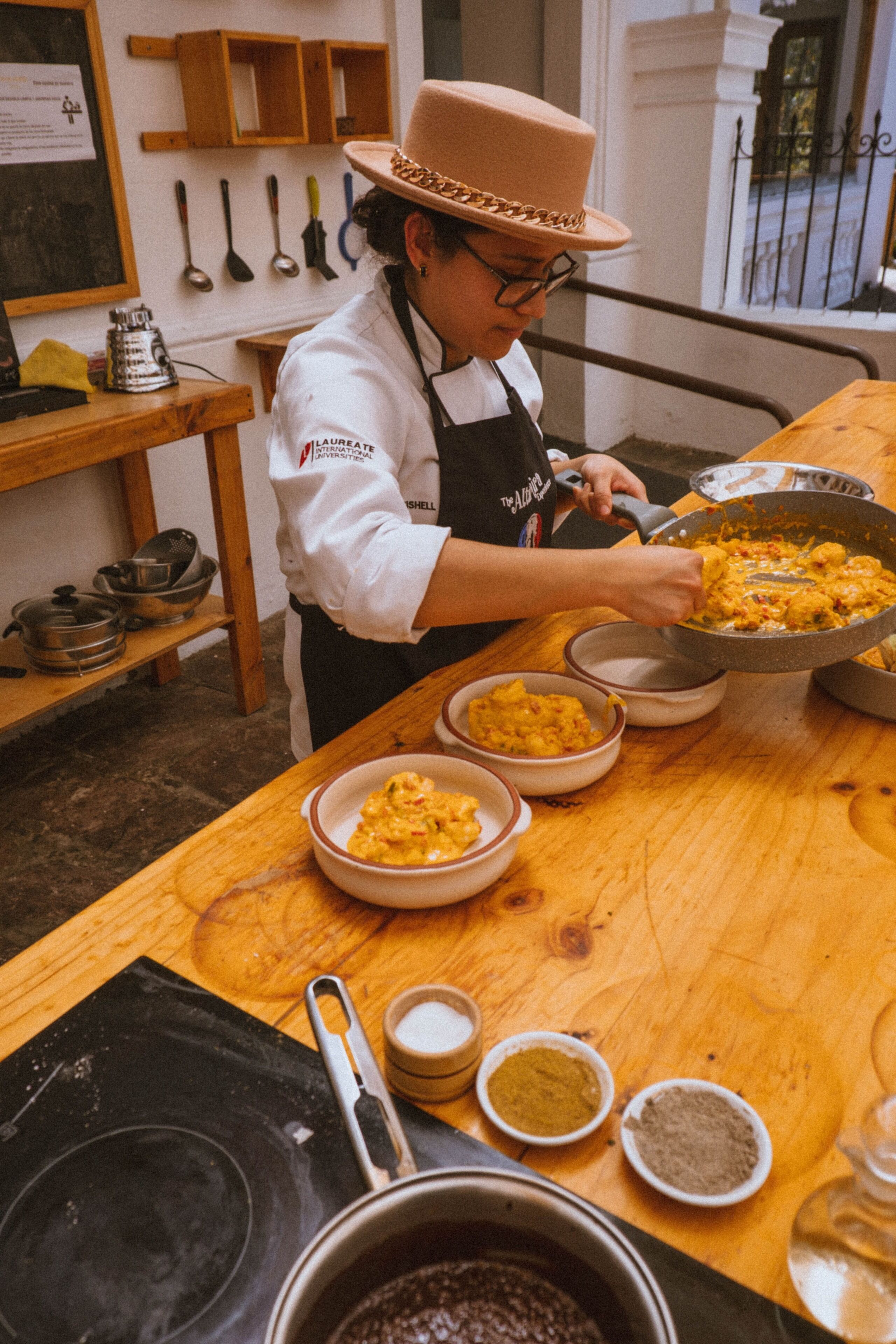A chef in a hat and apron preparing food in a kitchen with wooden tables and various cooking utensils.