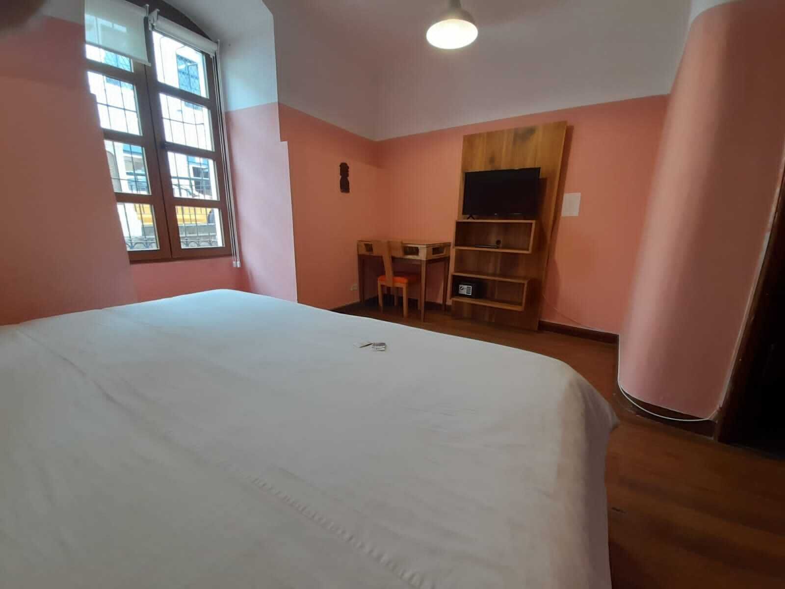 A bedroom with a large bed, a desk, a chair, and a TV shelf.