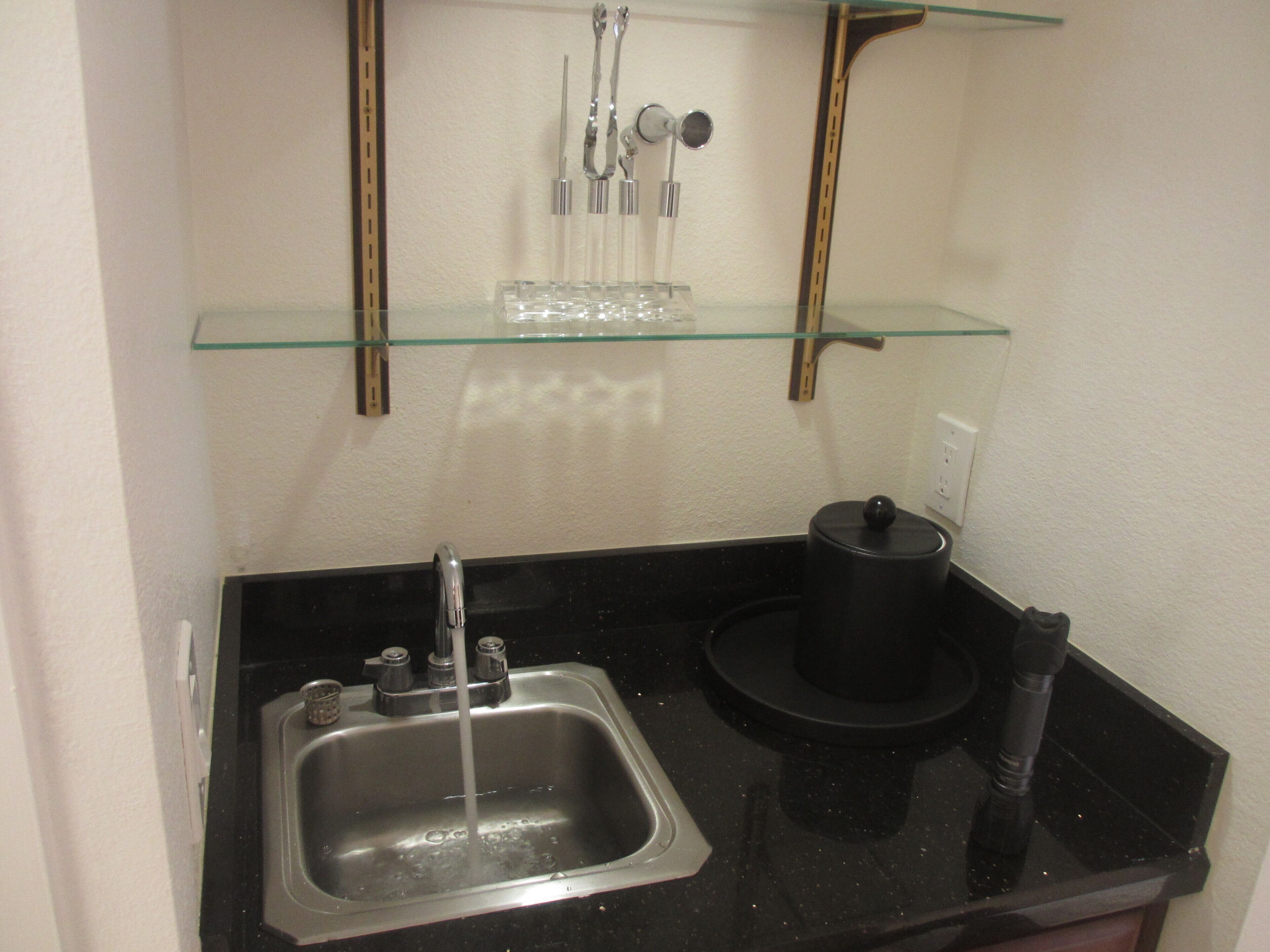 A small kitchen with a sink, a black countertop, and a glass shelf above the sink.