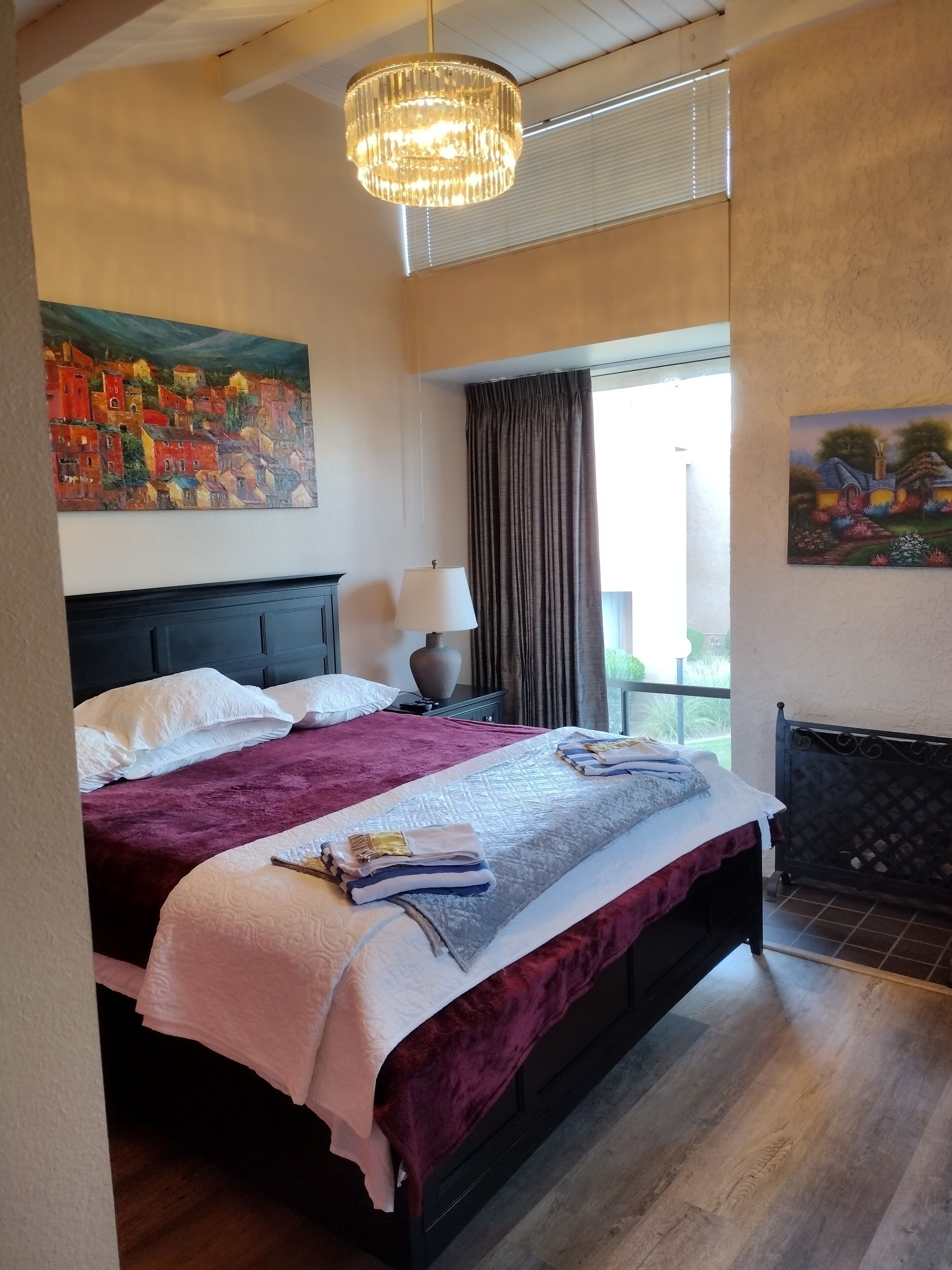 A bedroom with a large bed, a chandelier, two paintings, a window with curtains, and a radiator.