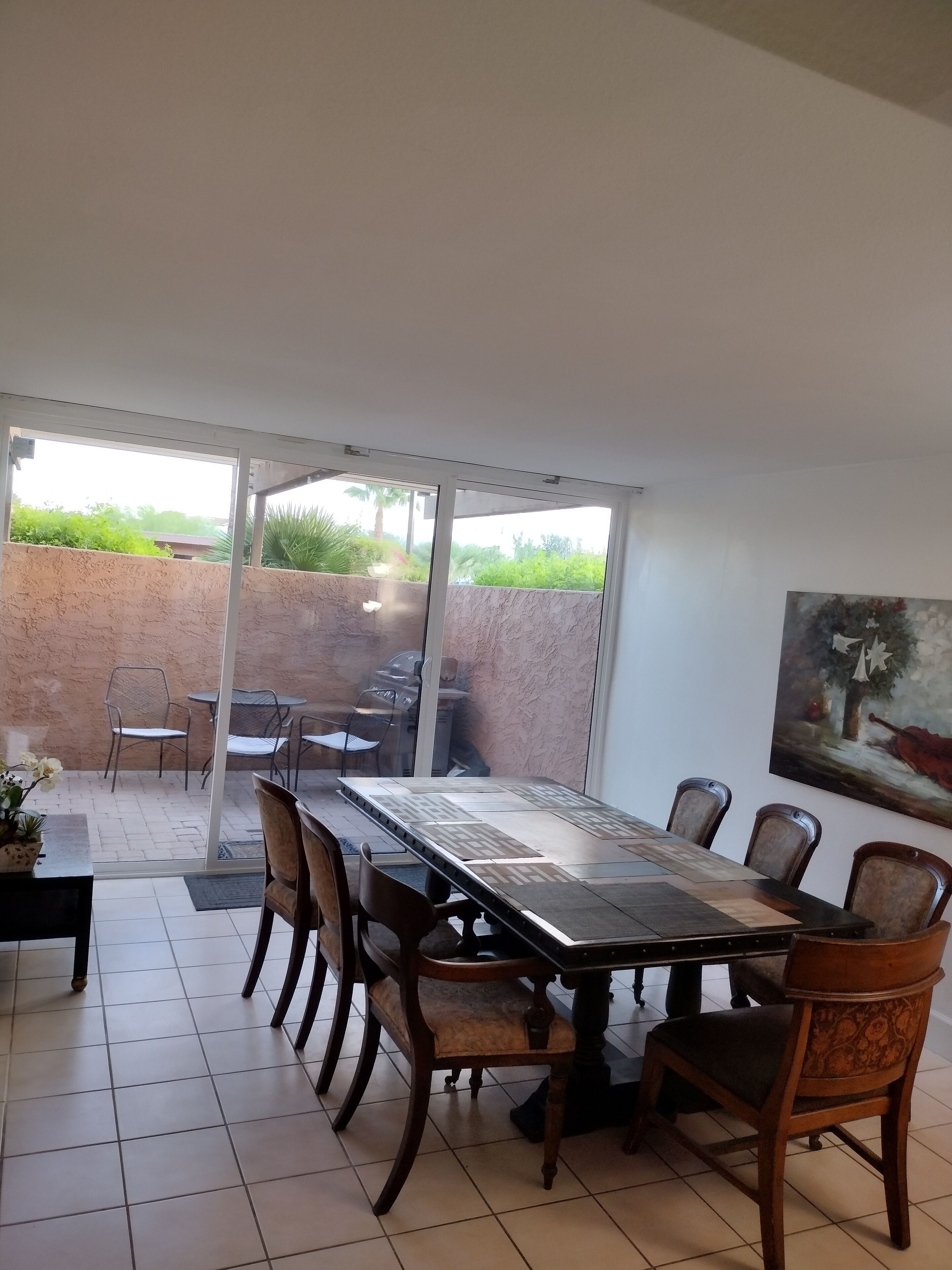 A dining area with a large table and chairs, a view of the outdoors through glass doors, and a painting on the wall.
