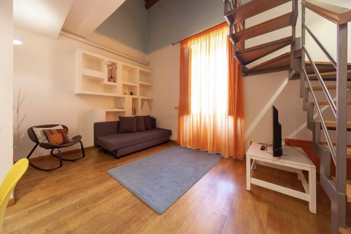 A living room with a sofa, a small table with a TV, a staircase, and a window with curtains.