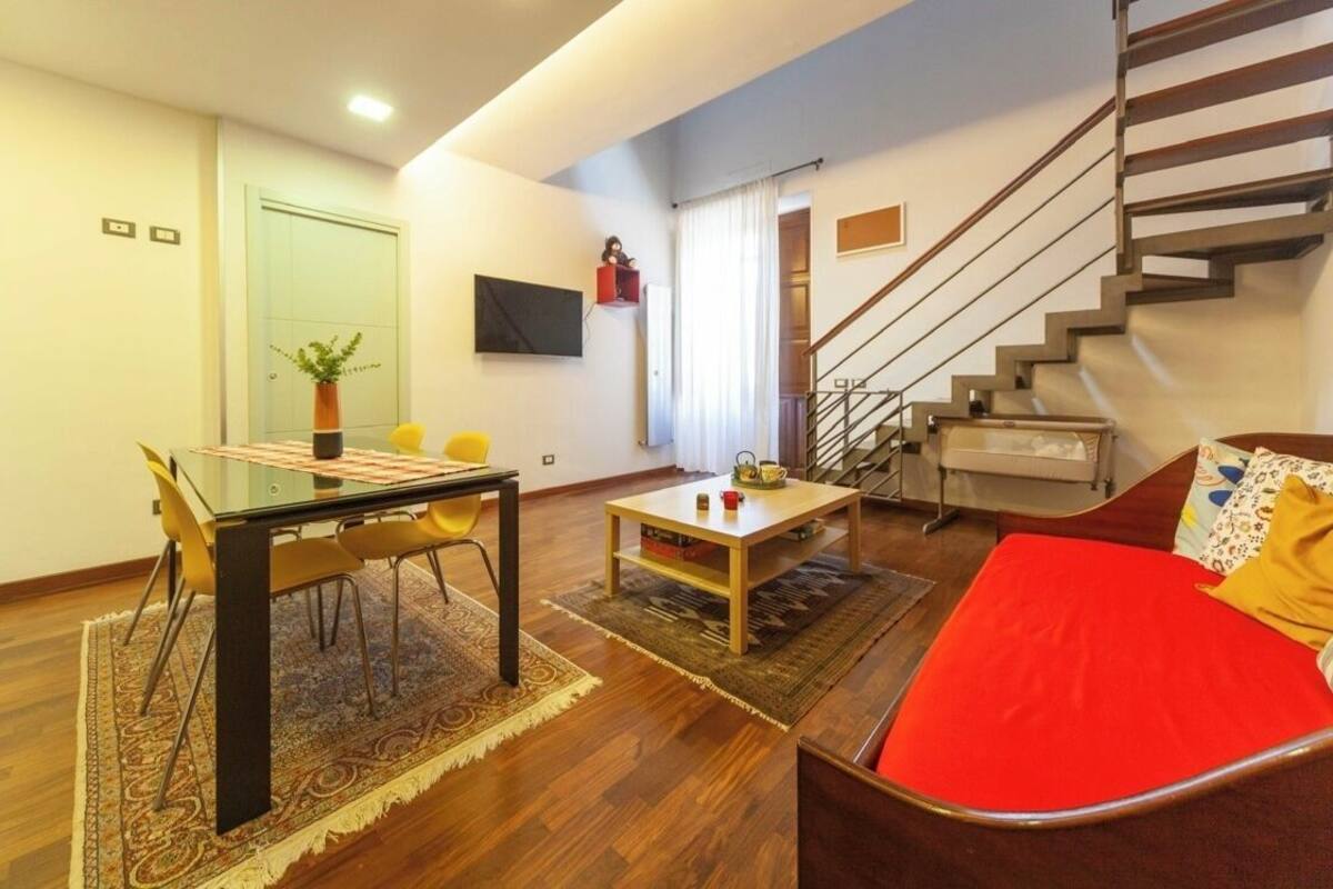 A modern living room with a glass dining table, yellow chairs, a red sofa, and a staircase.