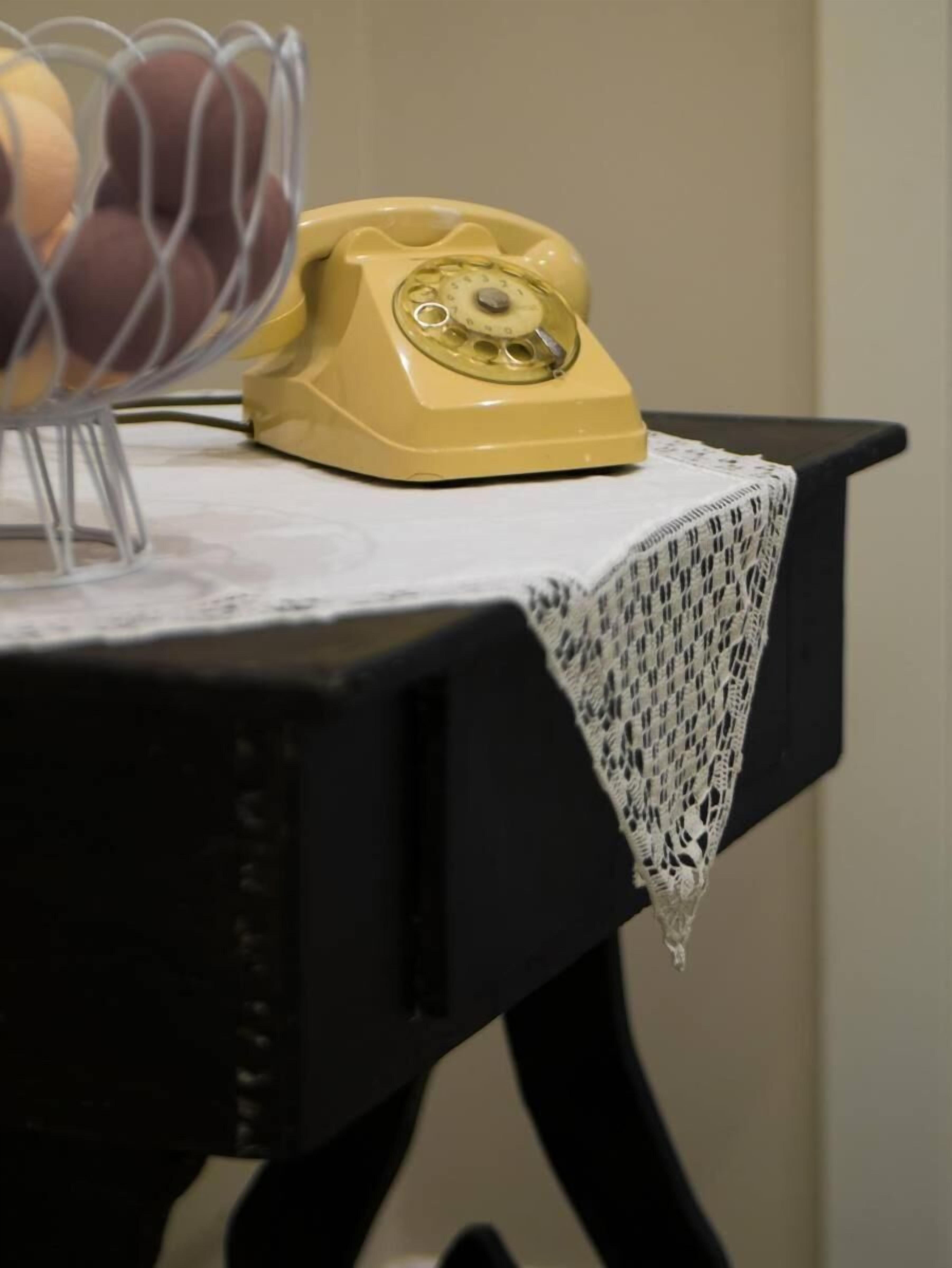 A yellow rotary phone on a table with a white lace tablecloth.