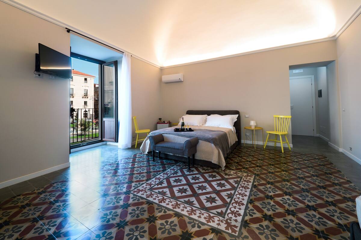 A bedroom with a bed, a patterned rug, a yellow chair, and a TV mounted on the wall.