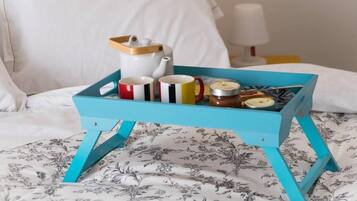 Uma bandeja azul-esverdeada com um bule branco, vários copos e potes sobre uma cama com roupa de cama floral.