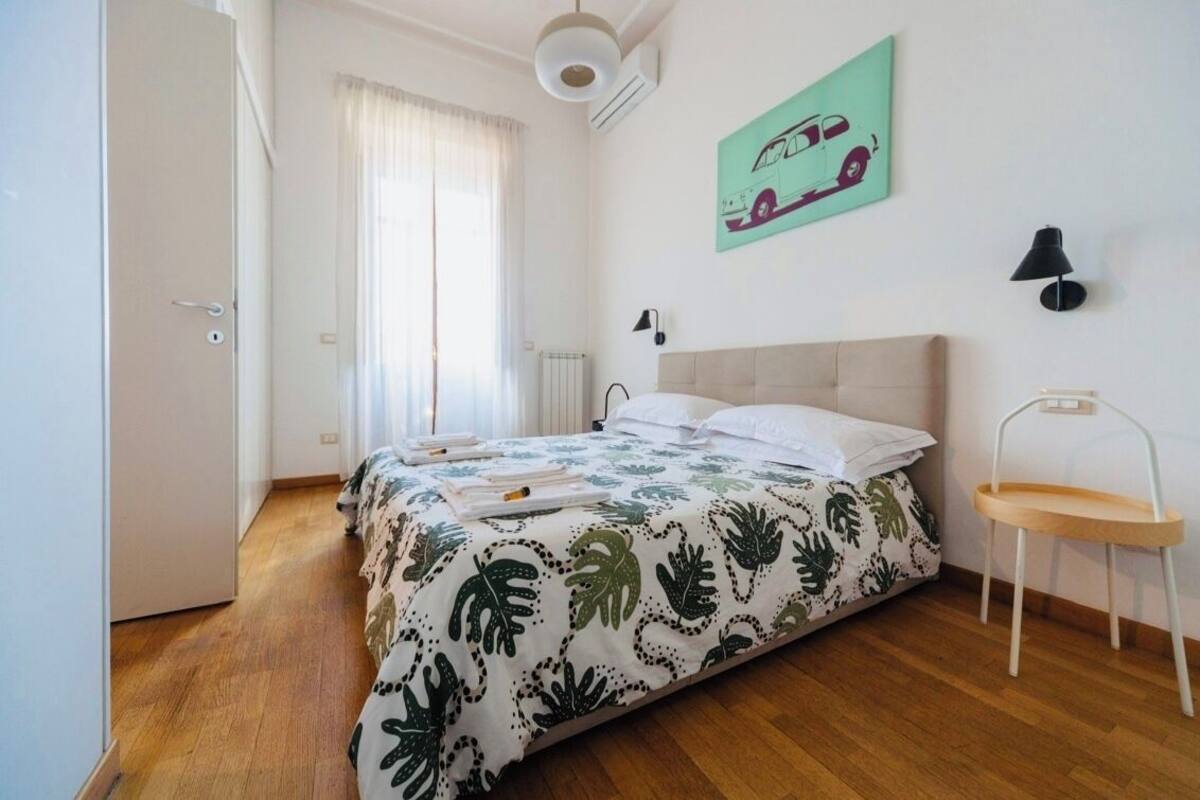 A bedroom with a bed, a radiator, a window with curtains, a wall-mounted lamp, and a door.