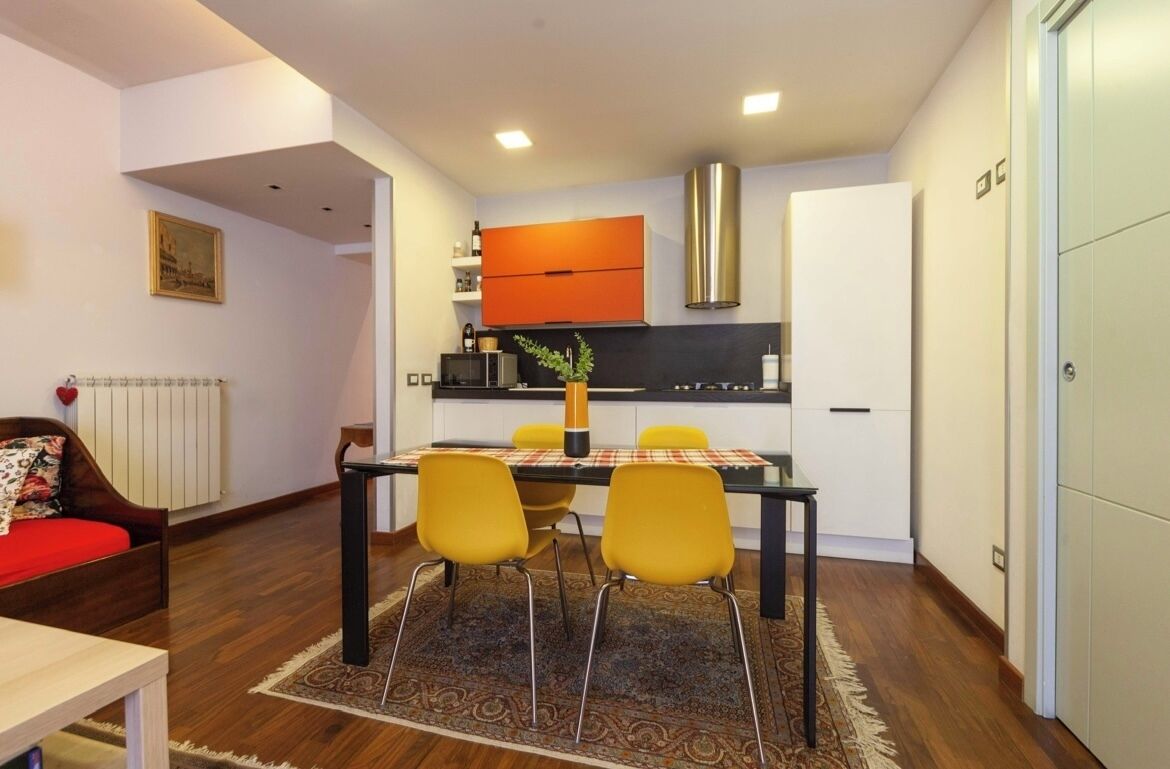 Un espace de vie compact comprenant un coin repas, un canapé, un radiateur et une cuisine avec des meubles orange et une hotte de cuisinière en acier inoxydable.