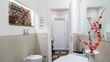 Un baño con inodoro, lavabo y zona de ducha. Hay un espejo, un jarrón con flores y un cuadro en la pared.