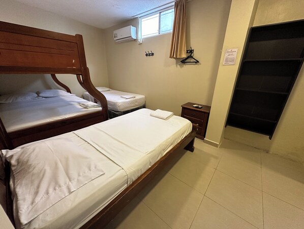 A bedroom with two wooden beds, a small bedside table, a wall-mounted air conditioner, a window with a curtain, and a door with a black handle. - Hostal Posada Del Sol (Guayaquil)