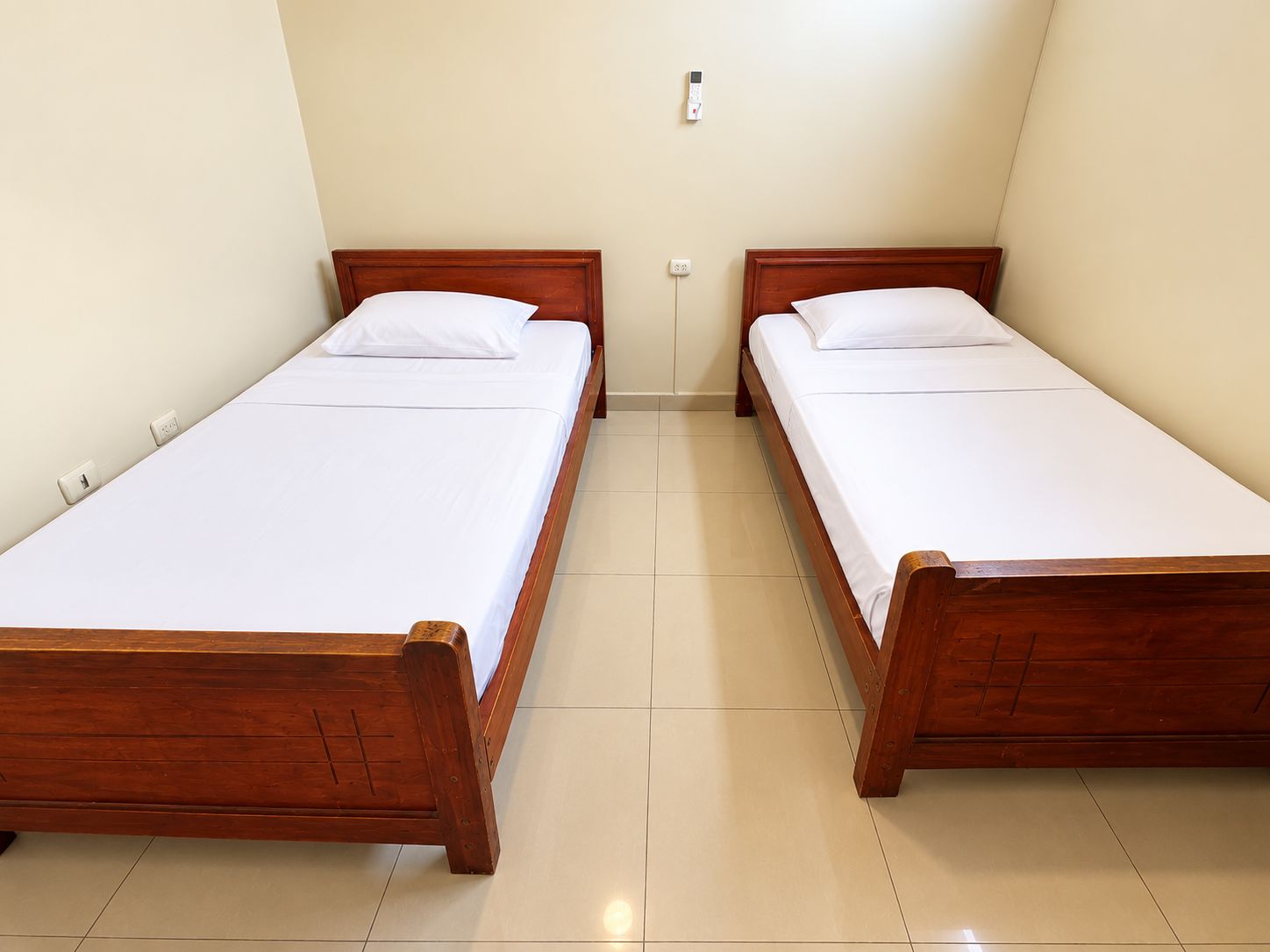 Dos camas de madera con ropa blanca en una habitación de paredes lisas y suelo de baldosas.