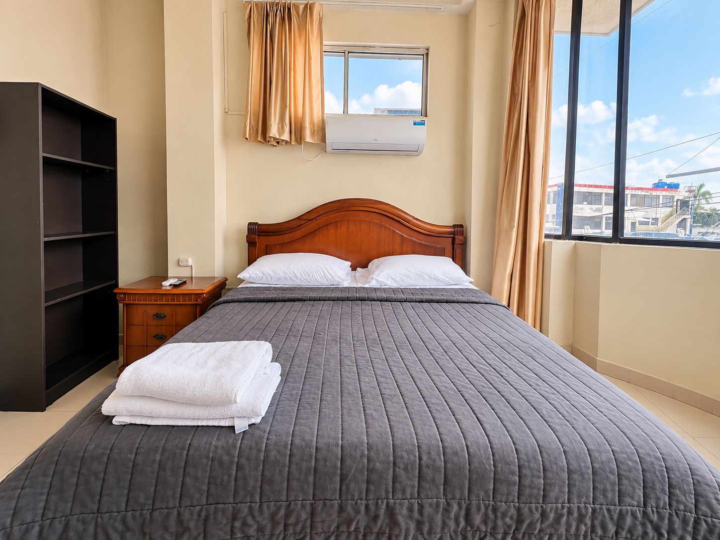 Habitación de hotel con cama, mesita de noche, armario, ventana con cortinas y unidad de aire acondicionado.