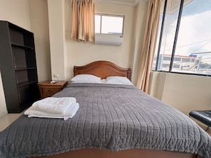 Een net opgemaakt bed met een grijze deken, witte kussens en een stapel witte handdoeken. Er is een houten nachtkastje met een lamp, een zwarte garderobe en een raam met gordijnen.