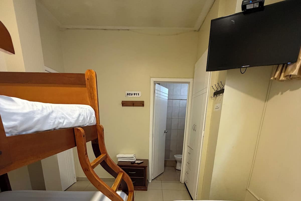 A small, narrow room with two wooden bunk beds, a small table with a chair, a television mounted on the wall, and a sign indicating the room number.