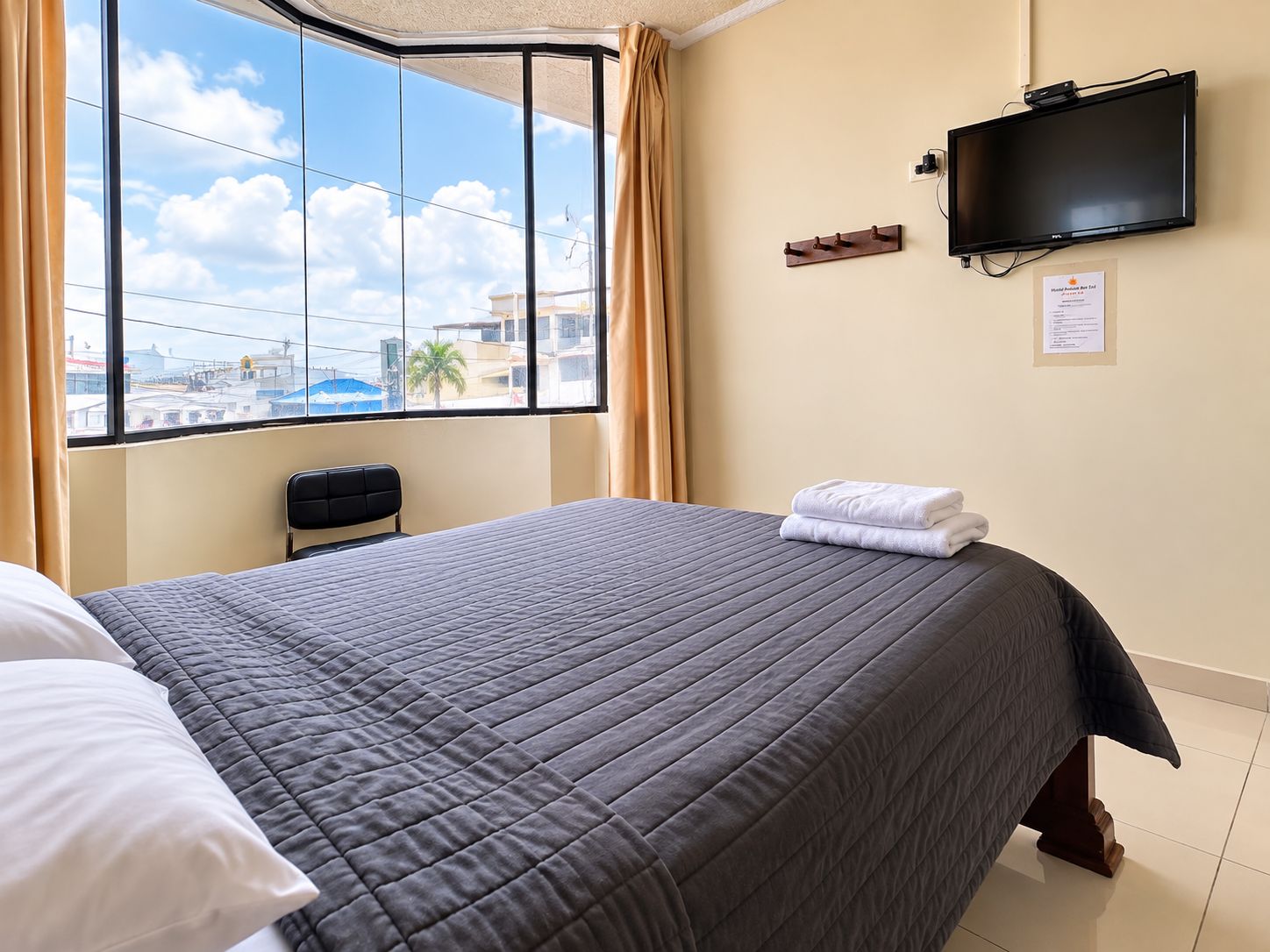 A hotel room with a large window, a bed with a grey quilt, a flat-screen TV mounted on the wall, and a chair.