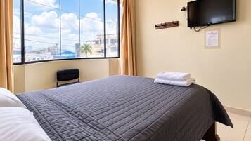 A hotel room with a large window, a bed with a grey quilt, a flat-screen TV mounted on the wall, and a chair.