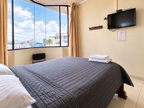 Habitación de hotel con un ventanal grande, cama con colcha gris, televisor de pantalla plana en la pared y una silla.