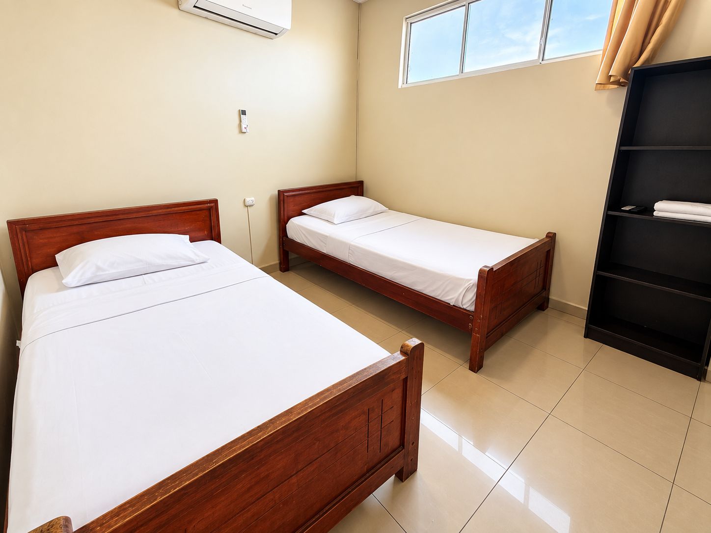 Una habitación con dos camas de madera, un estante negro y una ventana con cortinas.