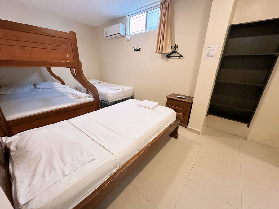 Quarto de hotel com duas camas de madeira, uma mesinha de cabeceira, ar condicionado fixado na parede, uma janela com cortina e uma porta com puxador preto.
