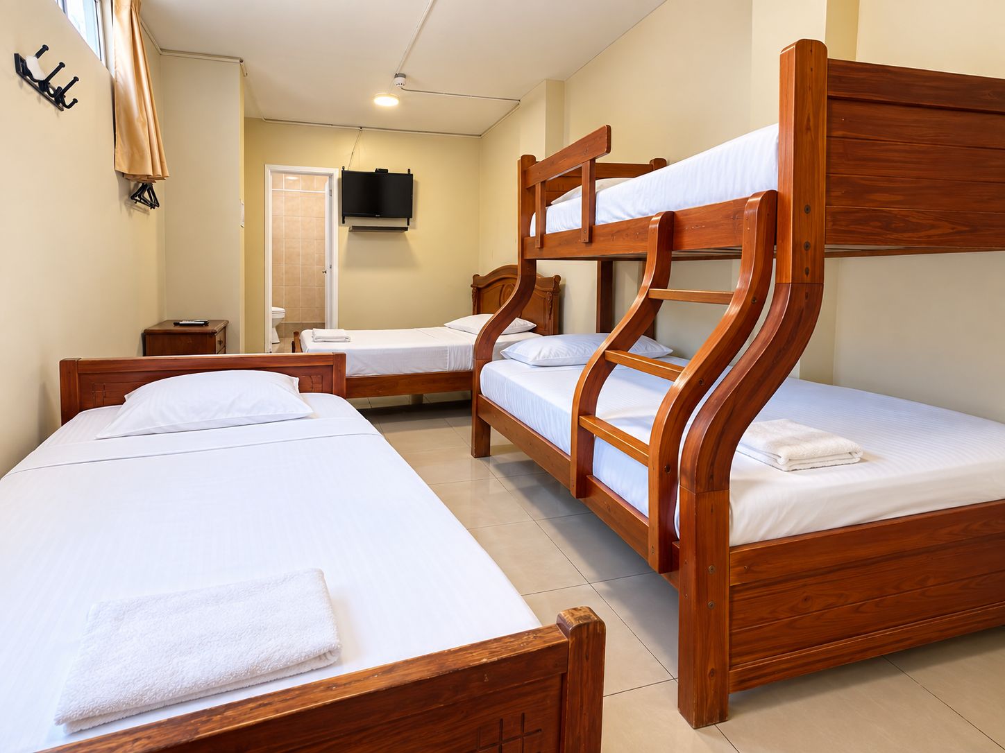 Una habitación de hotel con tres camas litera de madera, una mesita de noche pequeña, un televisor montado en la pared y una ventana con cortinas.